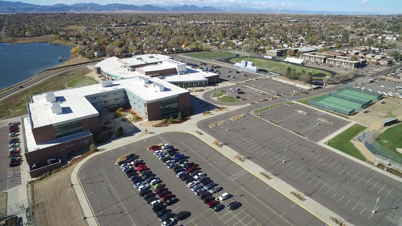 Opening aerial frame over Hidden Lake, Colorado, with a white-roofed commercial building complex, full parking rows, green tennis courts, a lake at left, distant mountains, and no visible people.