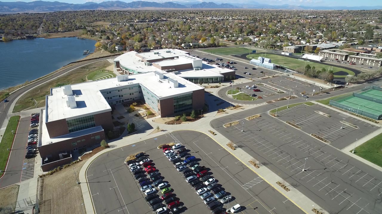 At 0:01, the Hidden Lake aerial view keeps the campus roofs central, with fuller car rows in the lower frame, empty parking spaces to the right, the lake to the left, and no visible people.