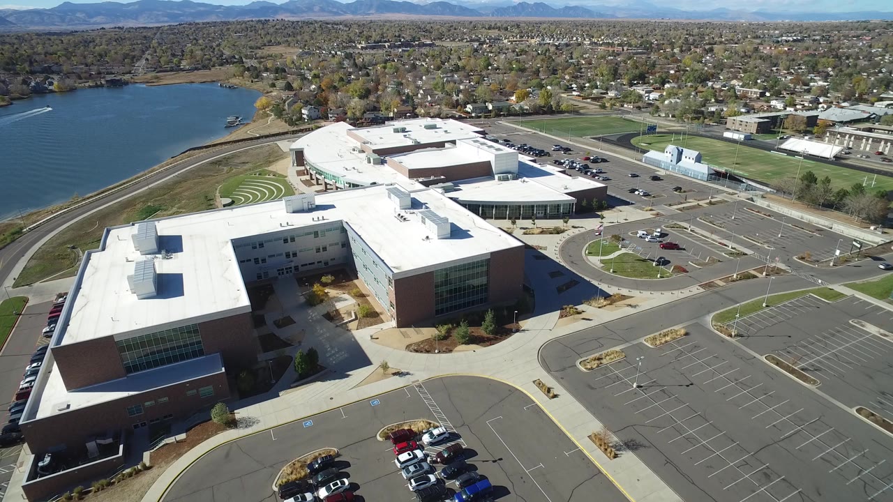 Opening aerial frame over Hidden Lake, Colorado shows white-roofed brick buildings, a lake to the left, paved parking lots, suburban trees, distant mountains, and no visible people.