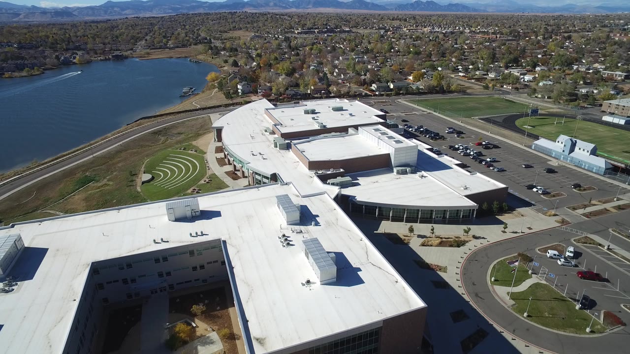 Drone view over a large white-roofed building beside a lake and neighborhood.