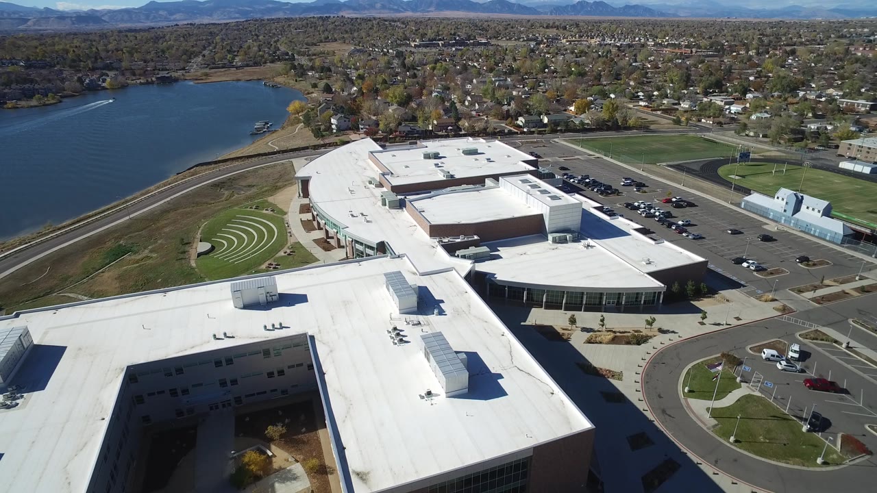 Second frame in Hidden Lake, Colorado, with the drone looking over the modern building roof toward the lake, parking areas, clustered houses, and no visible people.
