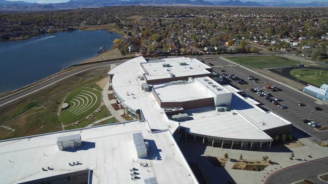 High aerial frame over Hidden Lake, Colorado showing a large white-roofed building complex, lake at left, parking lots at right, distant hills, and no visible people.