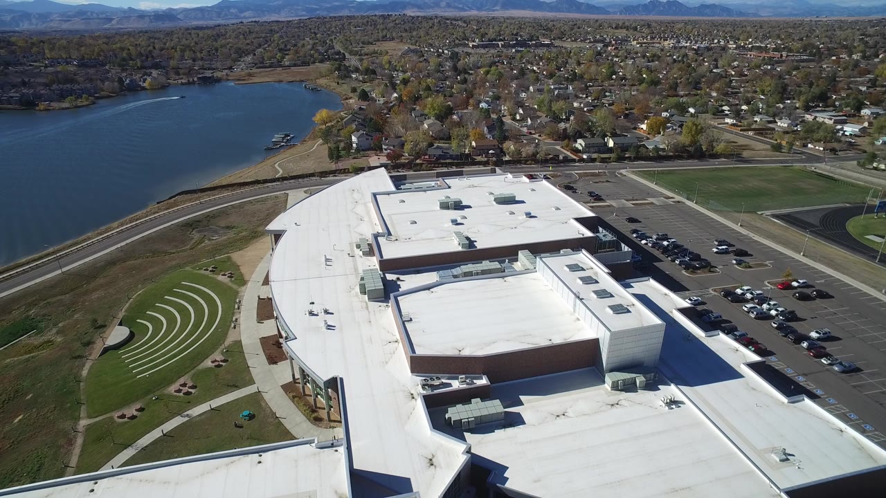 Aerial frame in Hidden Lake, Colorado showing a white-roofed commercial building, nearby lake, curved lawn terraces, parked cars, homes, distant hills, and no visible people.