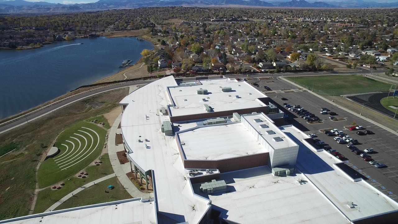 Aerial frame in Hidden Lake, Colorado with the white building roof filling the foreground, cars lined in the lot, the lake to the left, homes across the background, and no visible people.