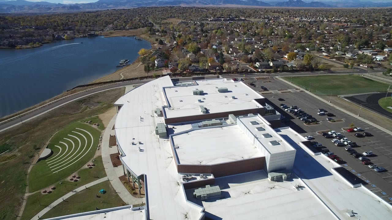 High aerial view of parking rows, a white commercial roof, lake edge, and neighborhood.