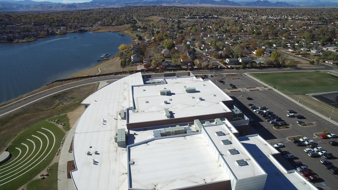 Aerial view over a white-roofed building beside a lake and neighborhood