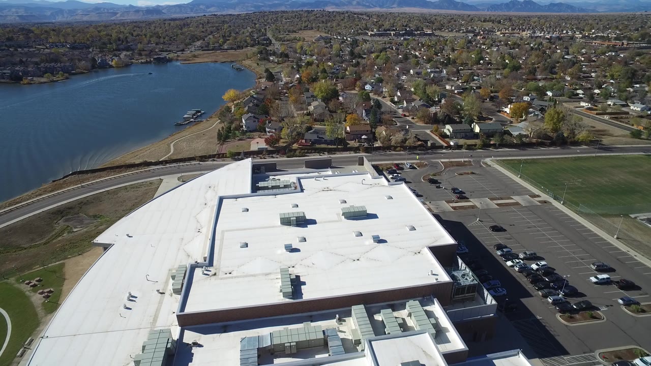 Aerial frame from Hidden Lake, Colorado showing the white roof lower center, a parking lot to the right, lake shoreline to the left, rows of homes beyond, and no visible people.