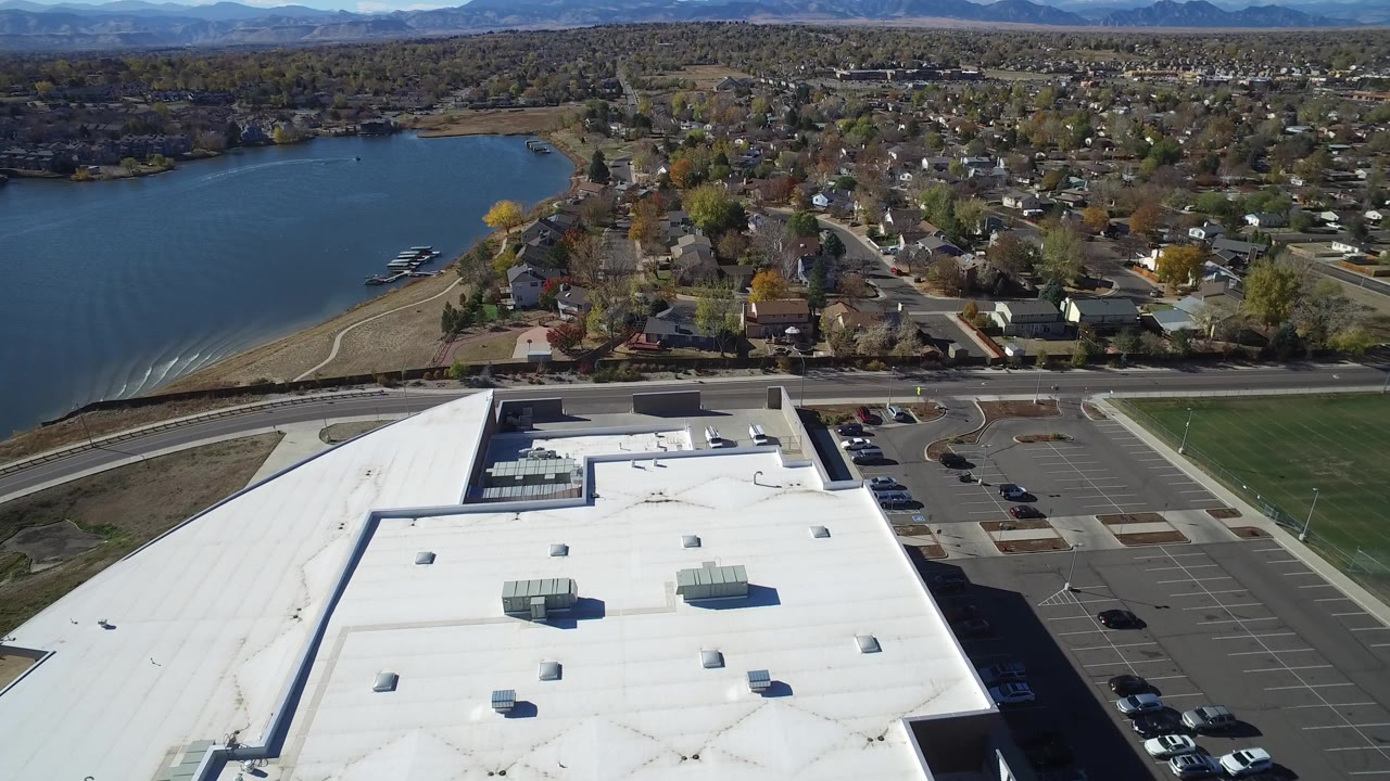 Aerial view over a white flat roof, parking lot, road, lake, and tree-lined homes.