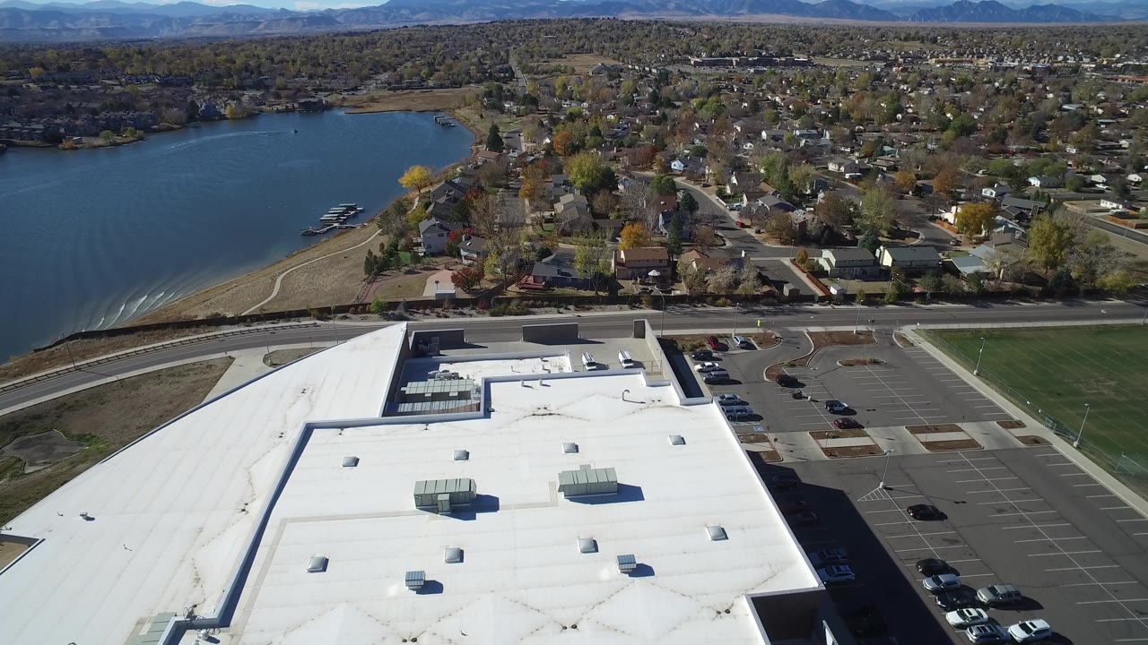 Aerial frame from Hidden Lake, Colorado with the white roof dominating the foreground, marked parking areas to the right, a road crossing the middle, and houses and lake beyond, with no visible people.