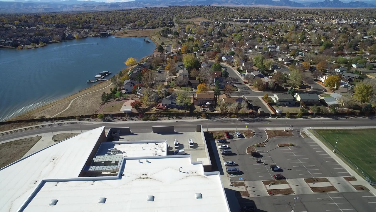 Opening aerial frame over Hidden Lake, Colorado, with blue lake water at left, residential streets in the center, a white flat roof and parking lots in the foreground, and no visible people.