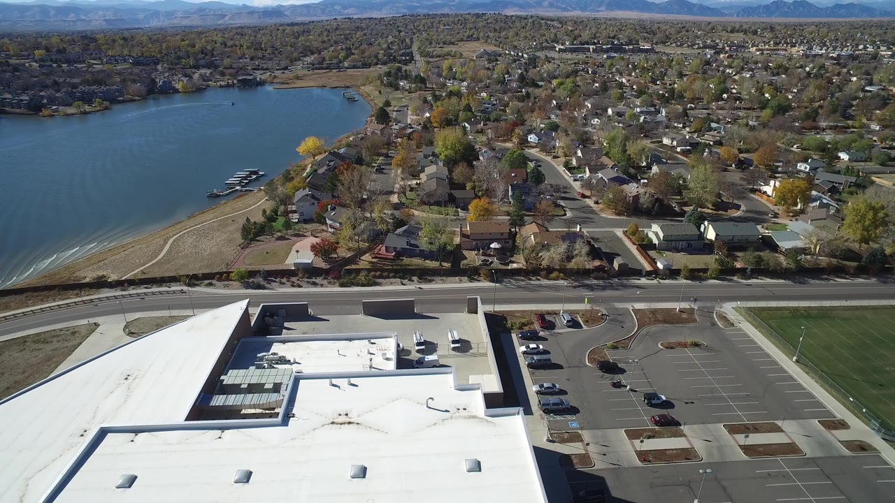 Aerial frame from Hidden Lake, Colorado showing the shoreline and docks on the left, a cluster of homes and trees ahead, a white roof below, parked cars in lots, and no visible people.