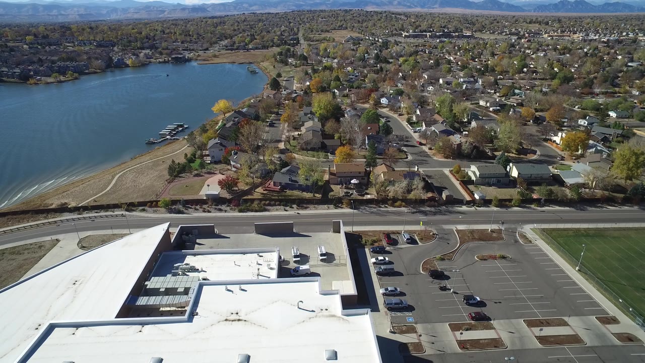Wide aerial of lakefront neighborhood with homes, trees, roads, roof, and parking lot.