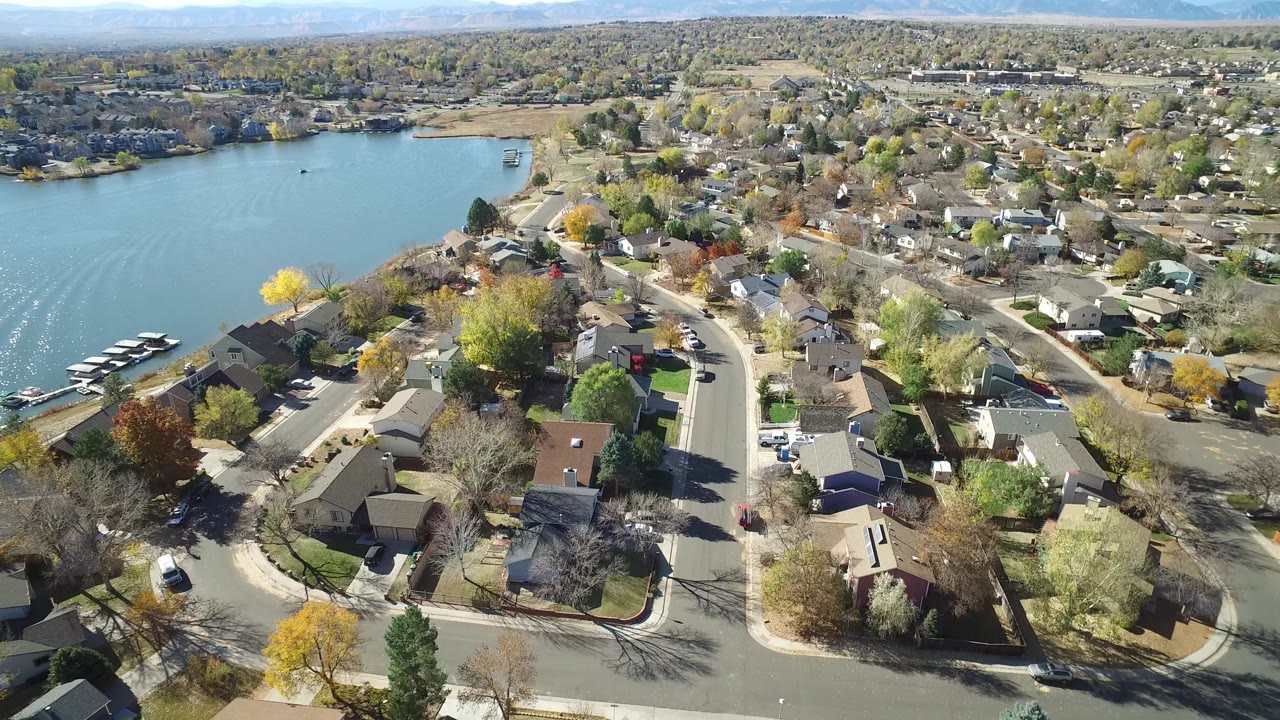 Opening aerial frame over Hidden Lake, Colorado, with blue water and docks on the left, curving streets and houses below, fall trees across the neighborhood, and no visible people.