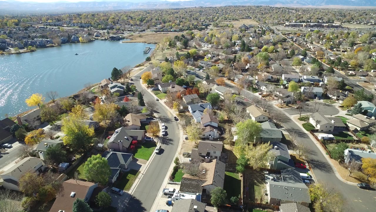 High aerial frame over Hidden Lake, Colorado homes with the lake on the left, curving streets below, autumn trees, rooftops, parked cars, and no visible people.