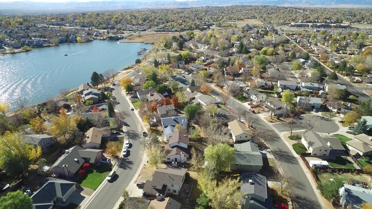 High aerial view in Hidden Lake, Colorado with the shimmering lake widening on the left, rooflines and backyards in the lower frame, gridded roads through the center, and no visible people.