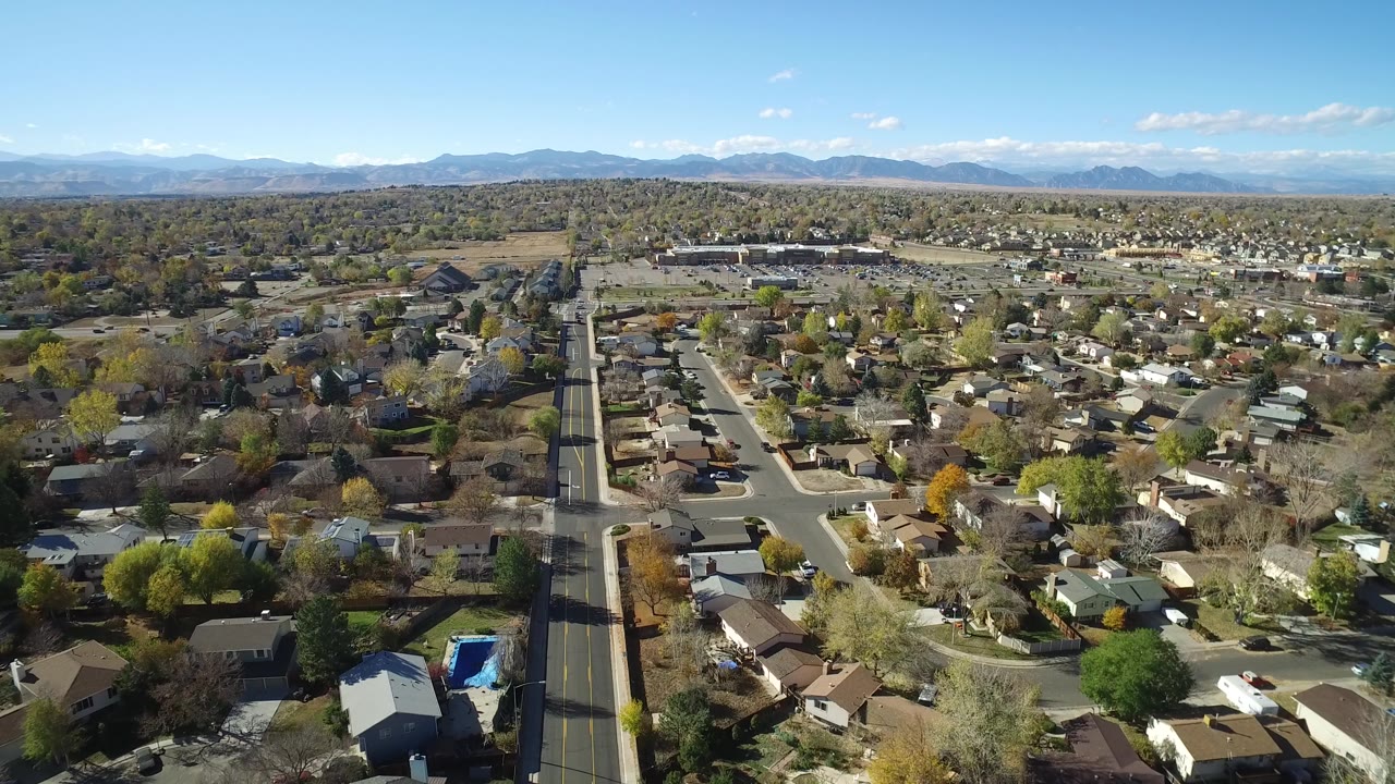 Aerial frame from Hidden Lake, Colorado showing a long road near the left side, residential blocks, autumn and green trees, a blue backyard pool, distant open land, and no visible people.