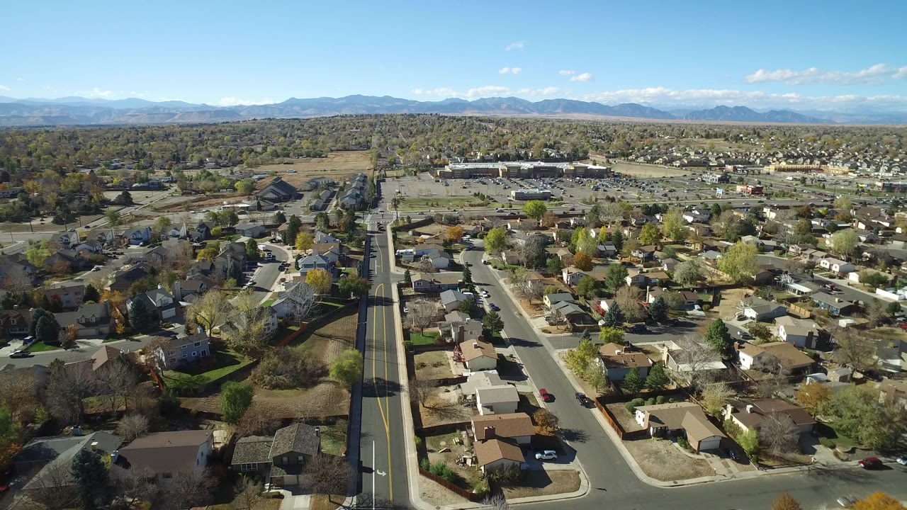Aerial frame over Hidden Lake, Colorado showing a long vertical road through homes, sparse traffic, brown and green yards, and mountains in the distance, no visible people.