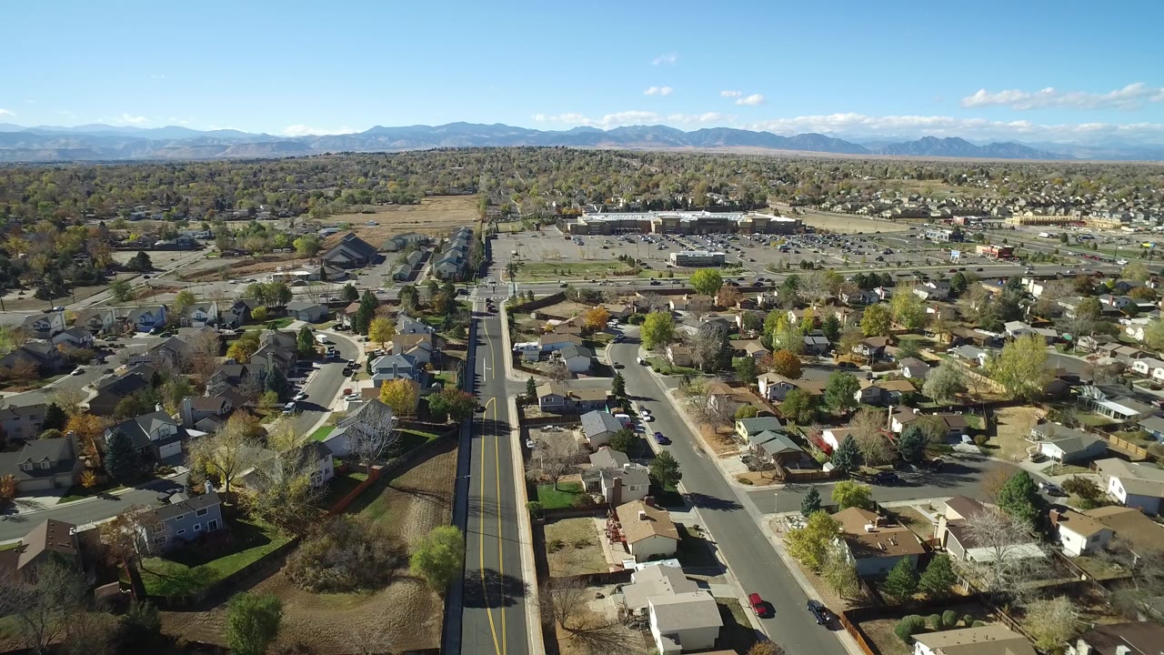Aerial frame over Hidden Lake, Colorado showing the central road slightly closer, single-family houses, parked cars, leafless and yellow trees, distant low buildings, mountains, and no visible people.