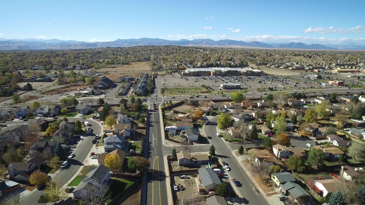 Opening aerial frame over Hidden Lake, Colorado, with curving residential streets in the foreground, a straight road through the center, distant mountains on the horizon, and no visible people.