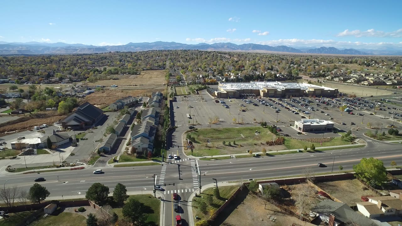 High aerial frame over Hidden Lake, Colorado showing a four-way intersection, crosswalks, nearby houses, a large parking lot, trees, distant mountains, and no visible people.