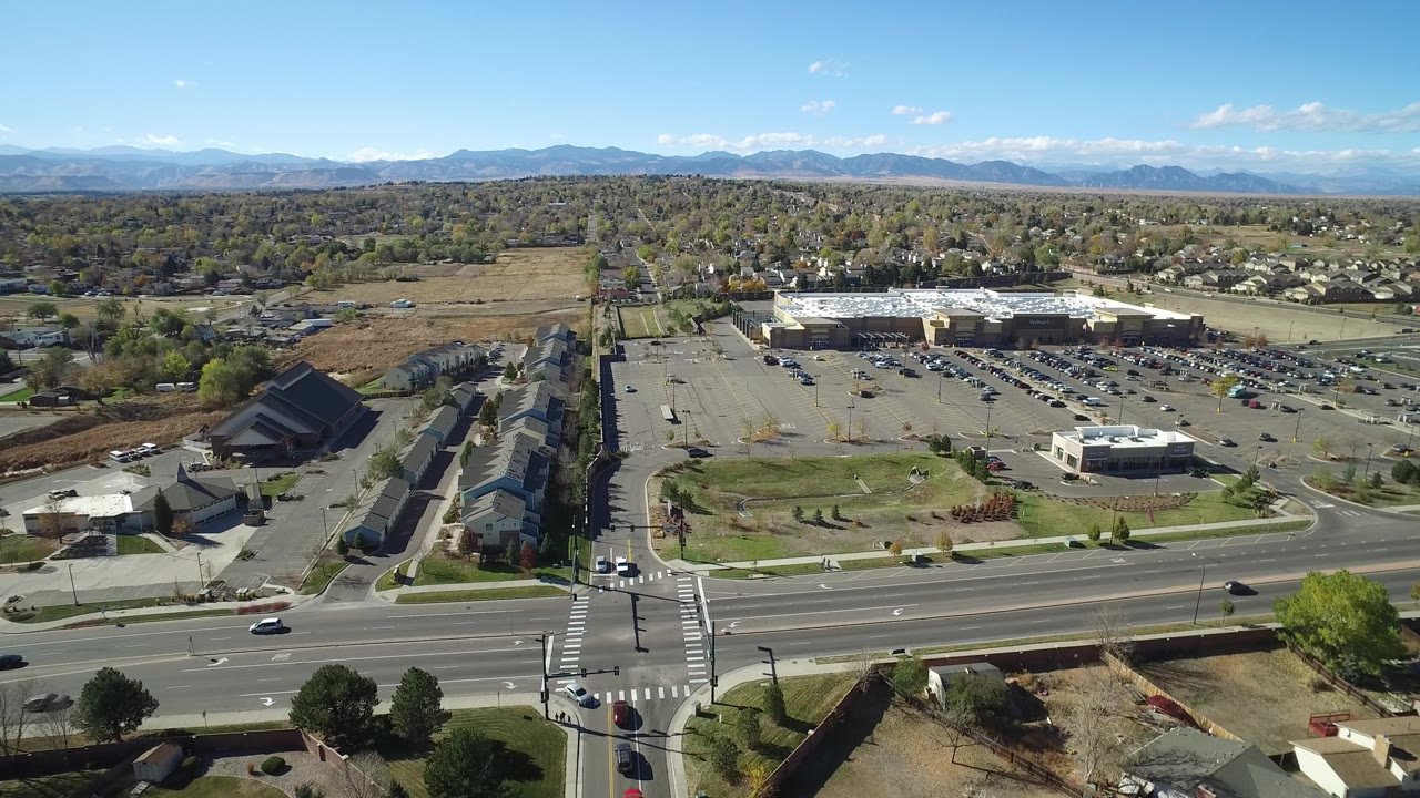 Drone-height view of roads, houses, parked cars, trees, and mountains under blue sky.