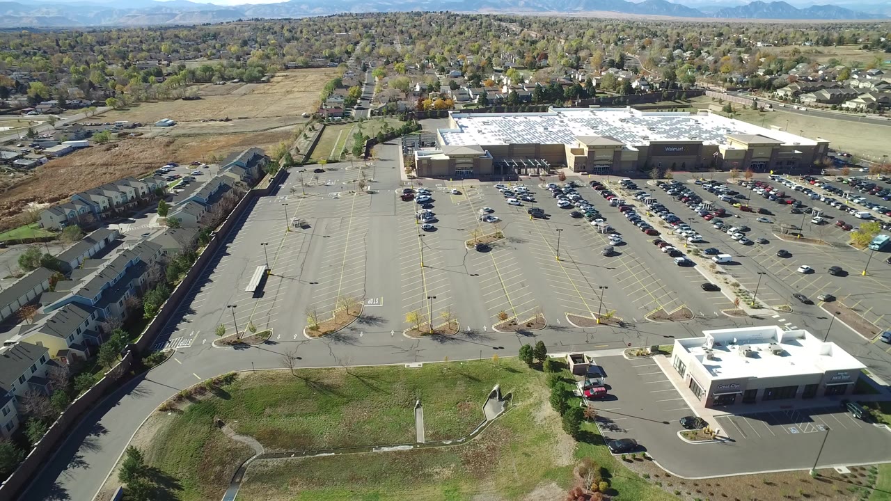 Aerial frame over Hidden Lake, Colorado shows a large retail building beyond a wide parking lot, a small white-roofed building in the foreground, apartments at left, distant homes, and no visible people.