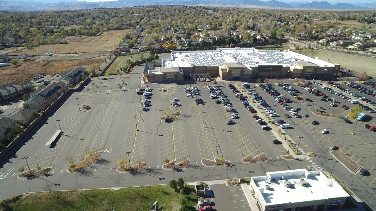 Aerial view in Hidden Lake, Colorado showing a wide commercial parking lot, rows of parked cars, a large retail building, nearby homes, distant hills, and no visible people.