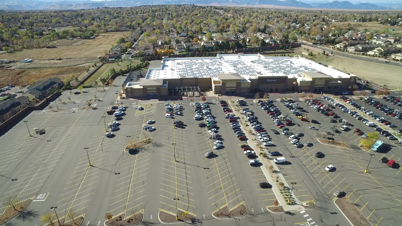 Second aerial frame in Hidden Lake, Colorado, looking over open parking rows, clusters of cars, light poles, the large retail building, nearby roads, homes, and no visible people.