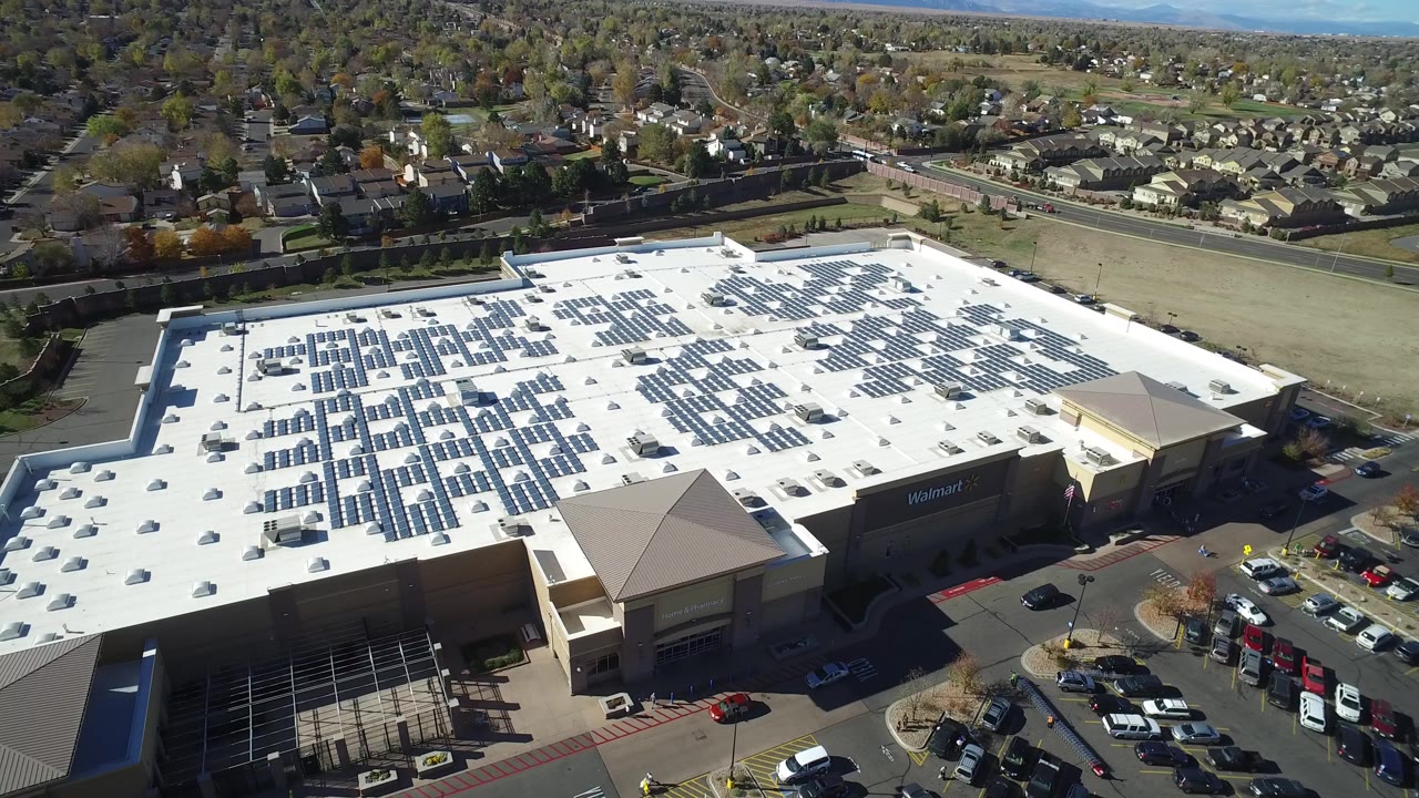 Oblique aerial frame in Hidden Lake, Colorado with dark solar panel rows across a white big-box roof, a Walmart sign on the facade, and homes in the background.