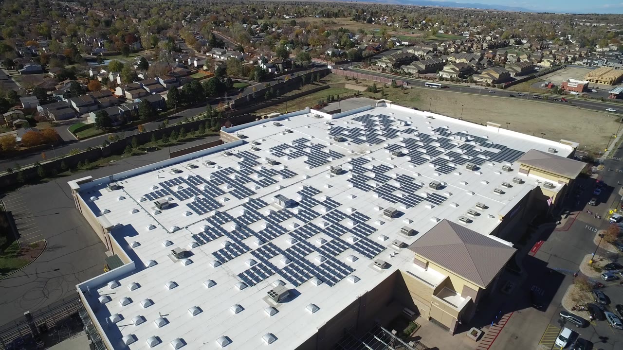 High aerial view in Hidden Lake, Colorado showing a large white flat roof with rows of solar panels, parking lots, roads, nearby homes, distant hills, and no visible people.