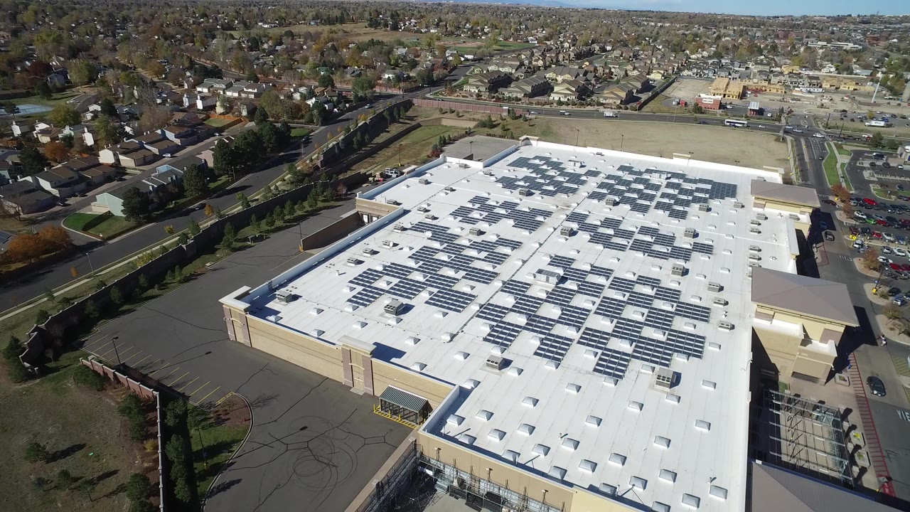 Aerial view in Hidden Lake, Colorado showing a large white-roofed commercial building with rooftop solar panels, adjacent parking areas, nearby roads, and no visible people.