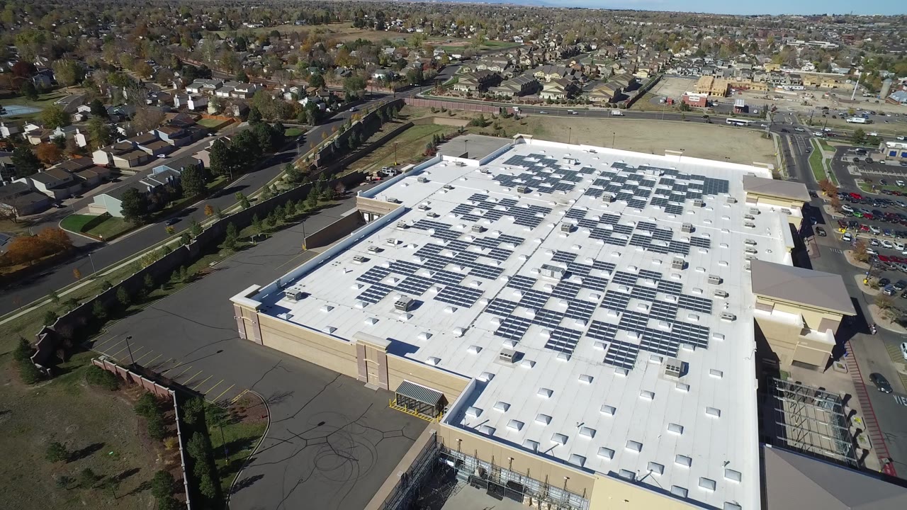 Top-down aerial of a broad white roof with solar panels and a parking lot beside it.
