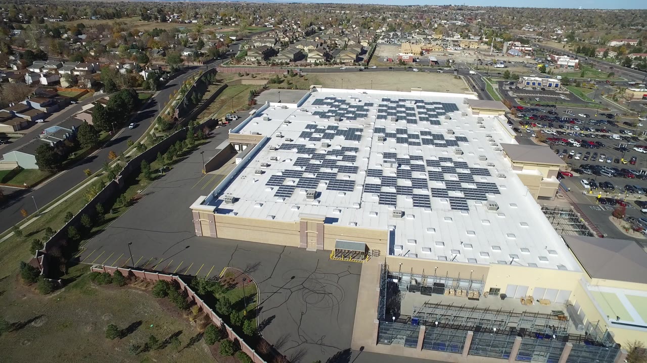 Aerial view over a large tan commercial building with a white roof and surrounding streets.