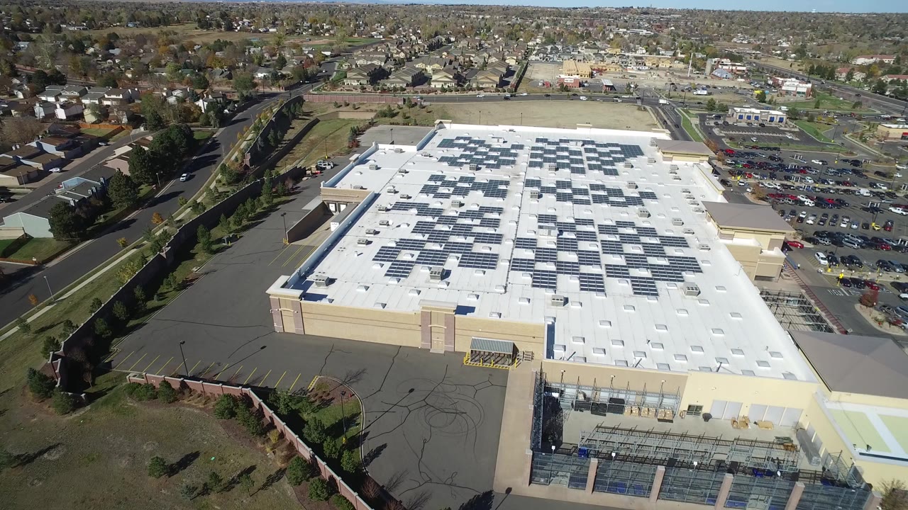 Second aerial frame in Hidden Lake, Colorado looking over the white roof and dark rooftop panel rows, with paved service areas, parking, roads, trees, and homes beyond with no visible people.