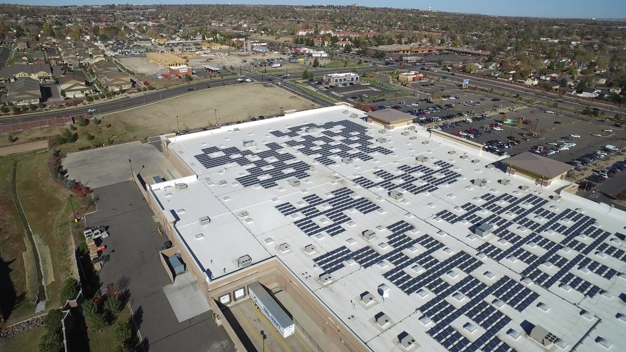 High oblique aerial view in Hidden Lake, Colorado showing a white commercial roof lined with dark solar panels, nearby parking lots, roads, and suburban blocks in the distance, with no visible people.