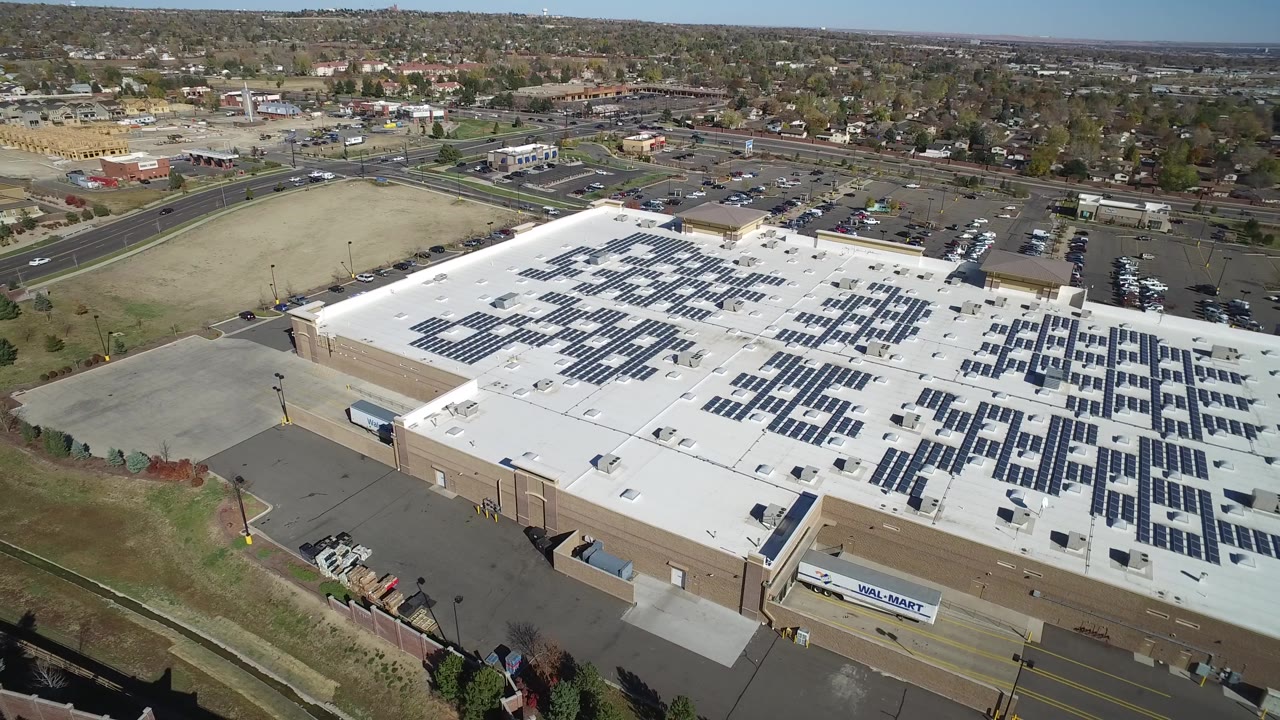 Aerial view one second into the clip showing more of the warehouse roof in Hidden Lake, Colorado, with solar panel grids, a vacant field, road lanes, parked vehicles, and no visible people.