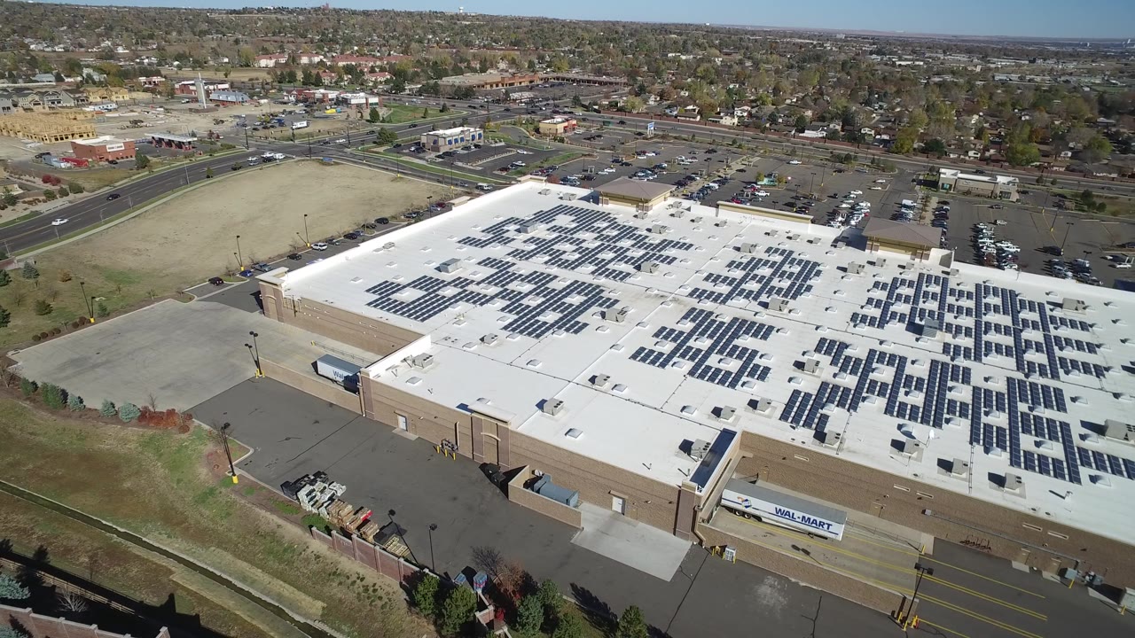 Oblique drone frame over the warehouse in Hidden Lake, Colorado, with the roof and solar rows pushed toward the right side, a parking lot and road grid behind it, and no visible people.