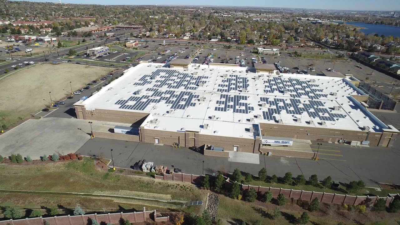 Second aerial frame near Hidden Lake, Colorado with the square white roof and solar panel grid centered more broadly against nearby parking lots, roads, suburban trees, distant water, and no visible people.