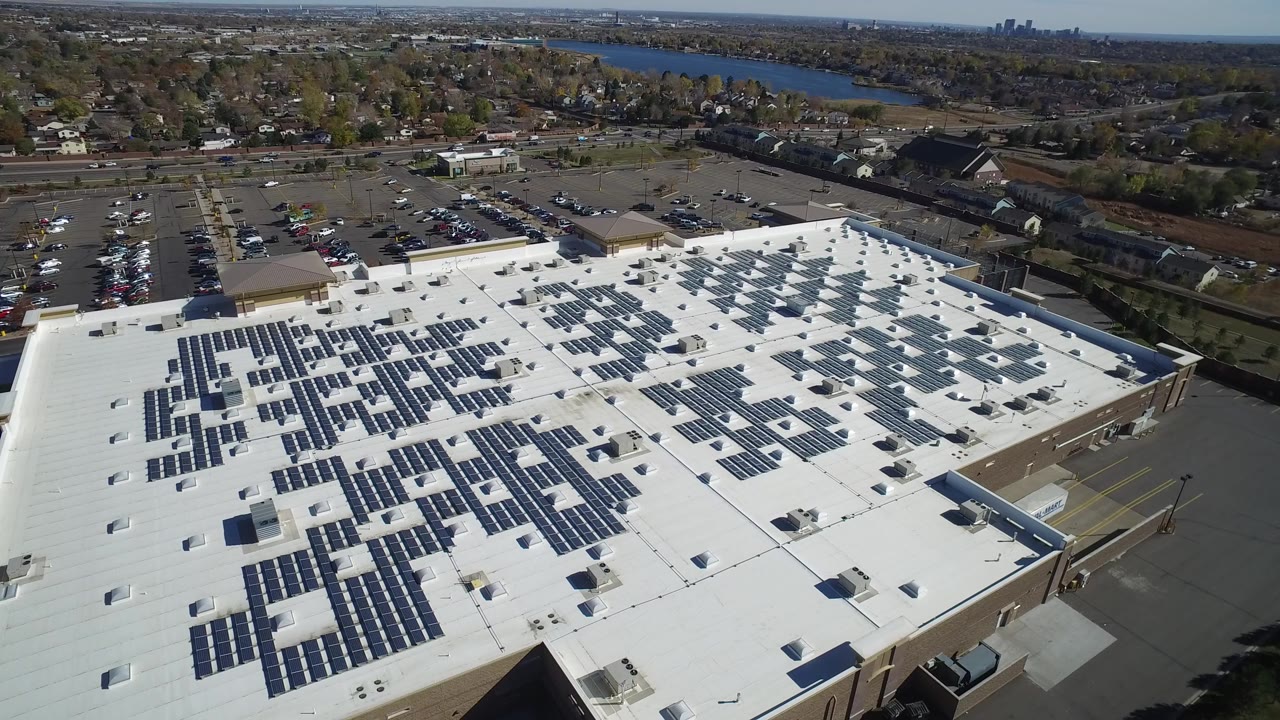 Aerial view of a white commercial roof with solar panels near parking lots and a lake.