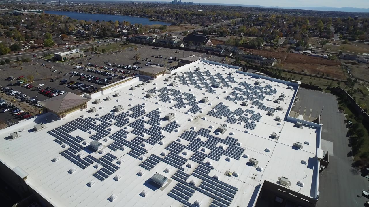Aerial frame from Hidden Lake, Colorado showing a large white commercial roof with dark solar-panel rows, parked cars, roads, neighborhoods, distant water, and no visible people.