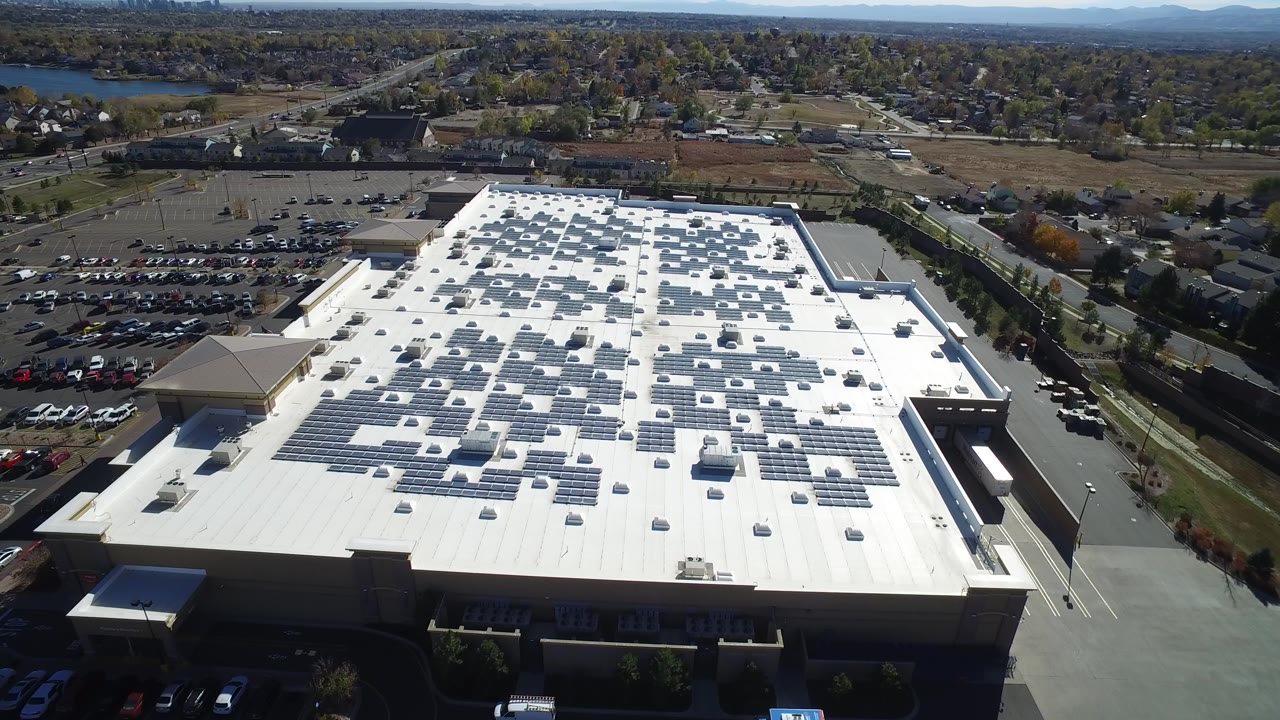 Aerial view of a large industrial roof with solar panels and parking lots.