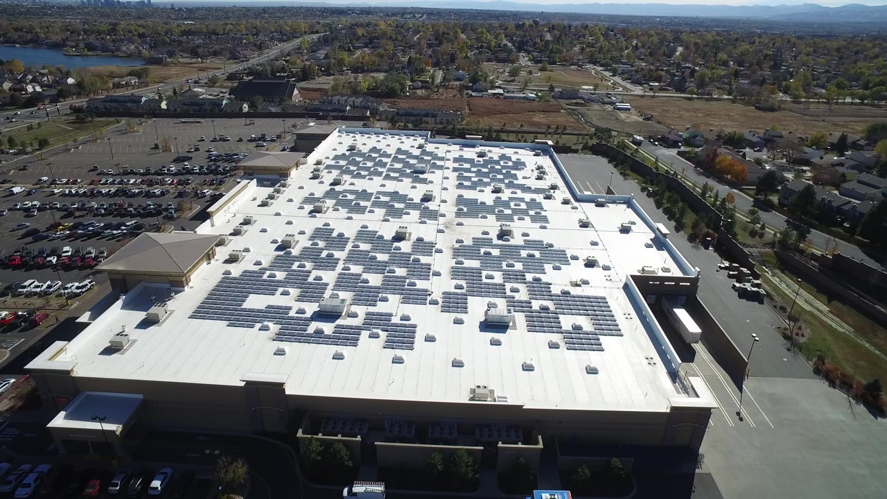 Aerial view in Hidden Lake, Colorado, with the white industrial roof centered, solar arrays spread across it, parking lots on both sides, suburban streets beyond, and no visible people.
