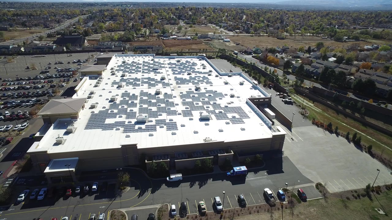 High aerial view in Hidden Lake, Colorado, looking over a large white-roofed commercial building covered with solar panels, with parking lots and roads around it and no visible people.
