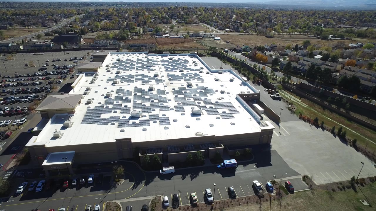 A slightly later aerial frame in Hidden Lake, Colorado, keeps the large solar-covered roof centered, with a row of parked cars along the front edge and no visible people.