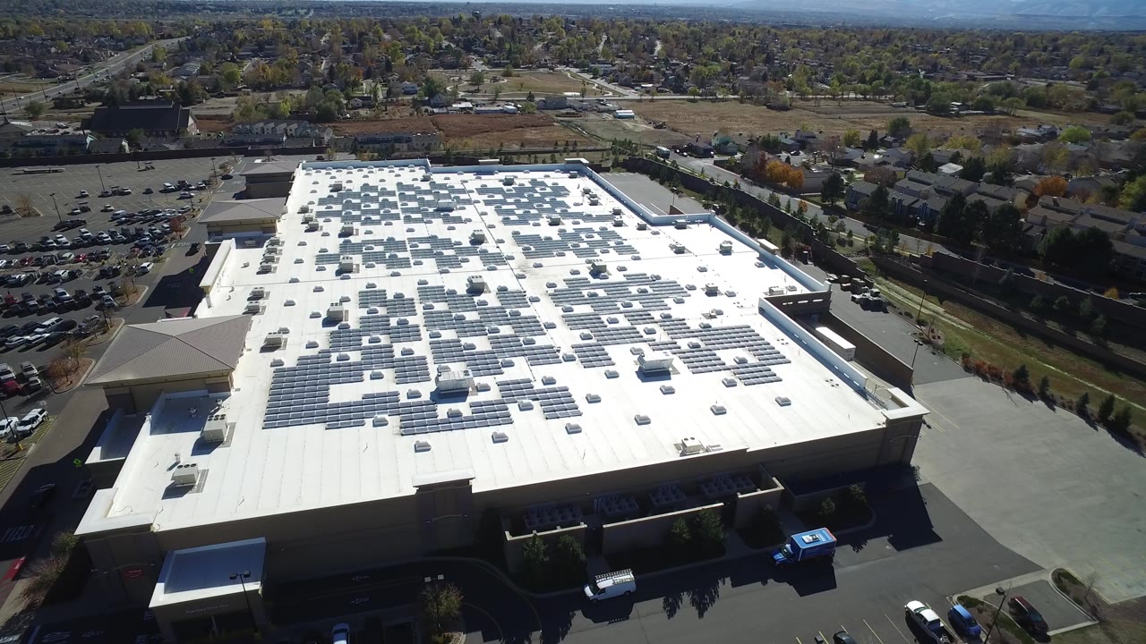 At 0:01, the Hidden Lake, Colorado aerial keeps the white roof centered, with solar panel rows, rooftop vents, a paved service area, cars, trees, and no visible people.