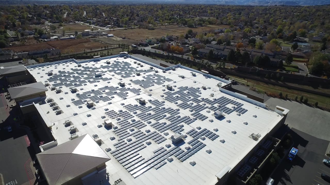 Aerial frame over Hidden Lake, Colorado, with the white roof and solar arrays centered, parking lanes along the side, suburban homes and open land in the background, and no visible people.