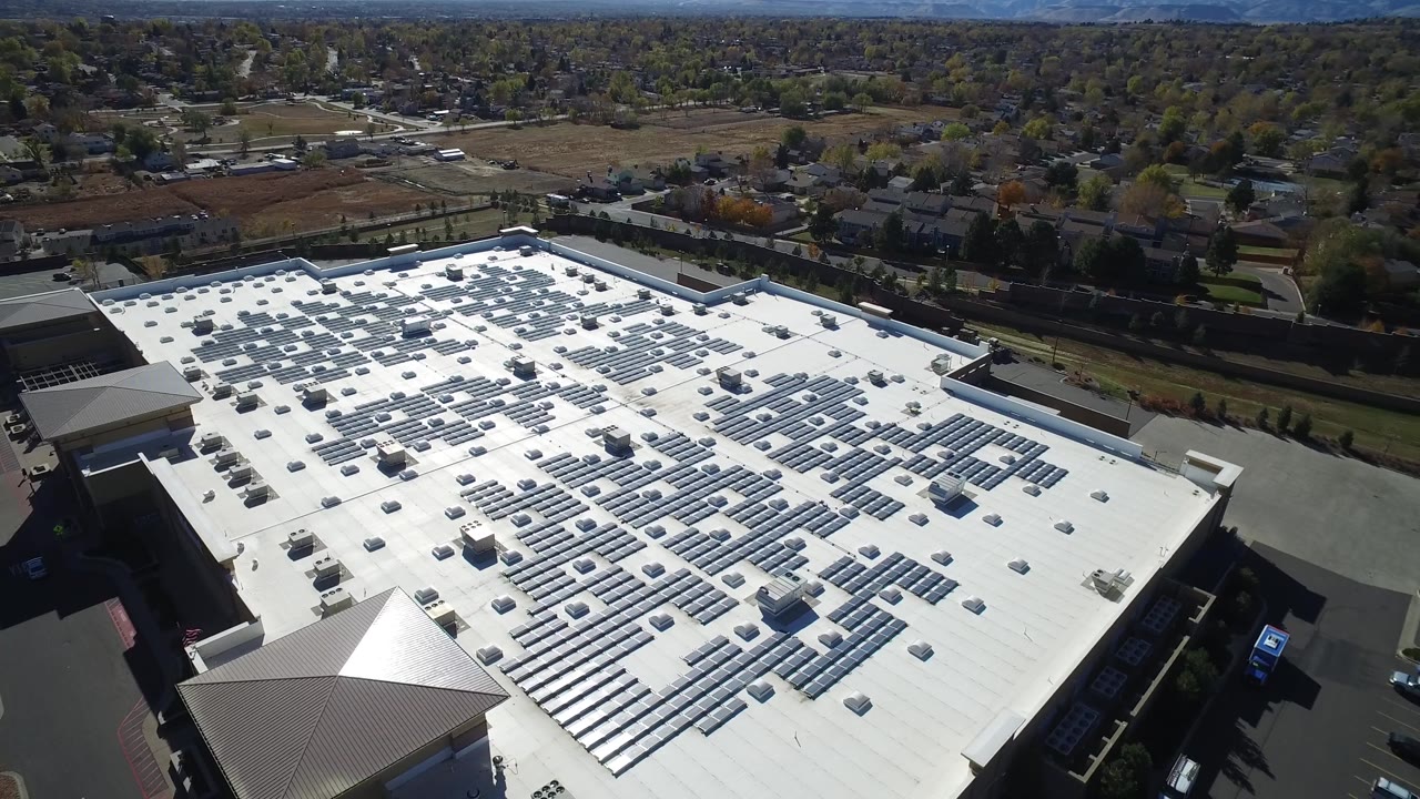 Oblique drone view in Hidden Lake, Colorado, showing a broad commercial rooftop with solar panels and mechanical units, a driveway below, residential blocks beyond, and no visible people.