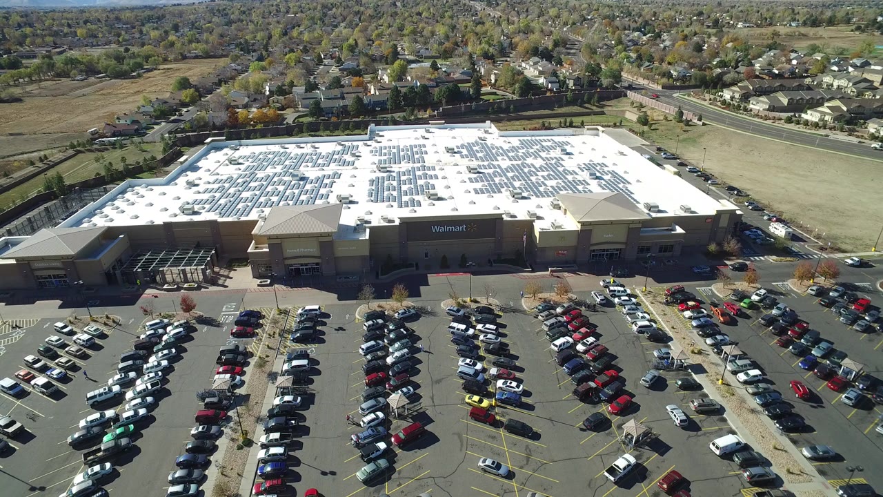 Aerial frame in Hidden Lake, Colorado showing a Walmart building with a white solar-panel roof, rows of parked cars in the foreground, roads and homes beyond, and no visible people.