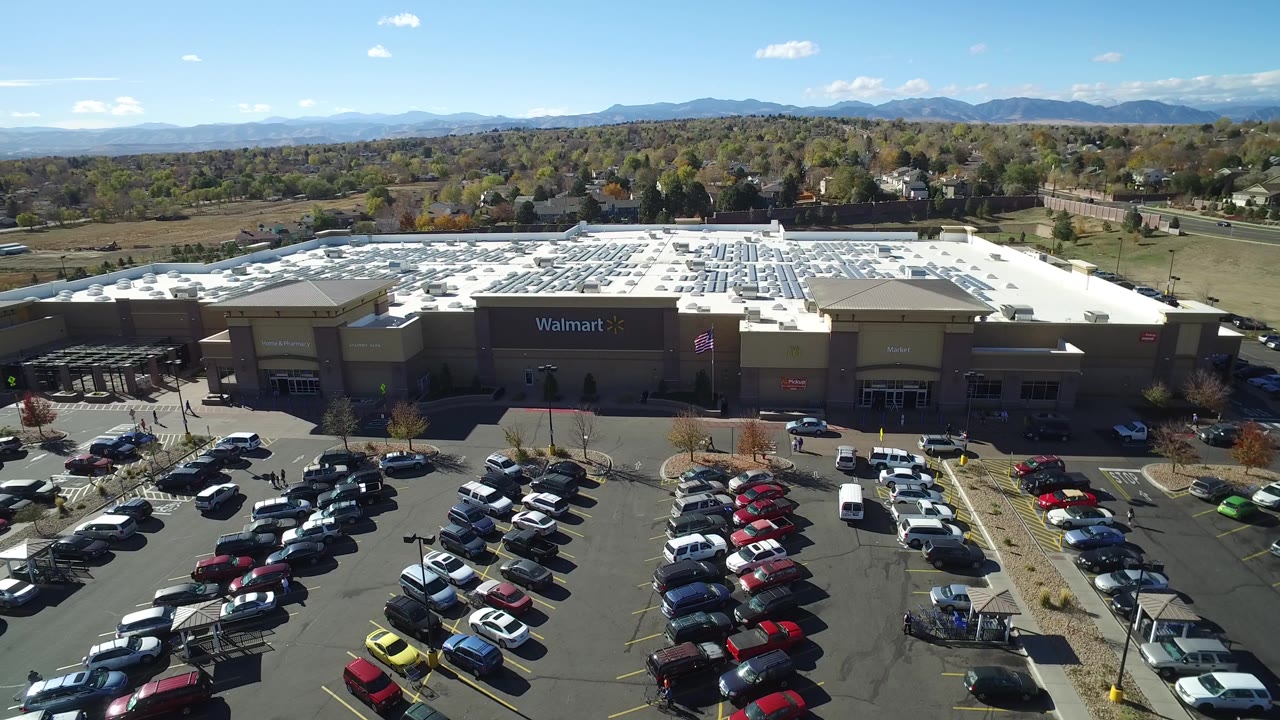 Aerial frame in Hidden Lake, Colorado showing the Walmart sign centered on the facade, car rows below, rooftop equipment catching sunlight, distant houses and trees, and very small people near the store entrance.