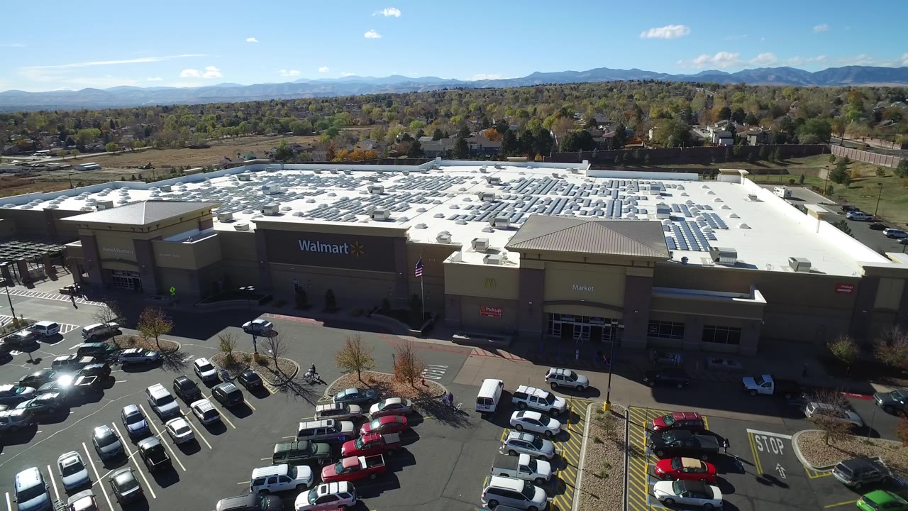 Aerial view in Hidden Lake, Colorado with the Walmart sign centered on the building, rooftop solar panels catching sun, parked cars below, and no visible people.